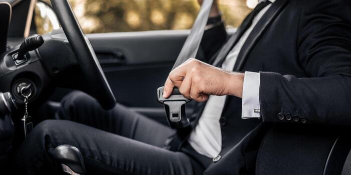 A person in a dark suit seated in a car, drawing the shoulder belt across before setting off.