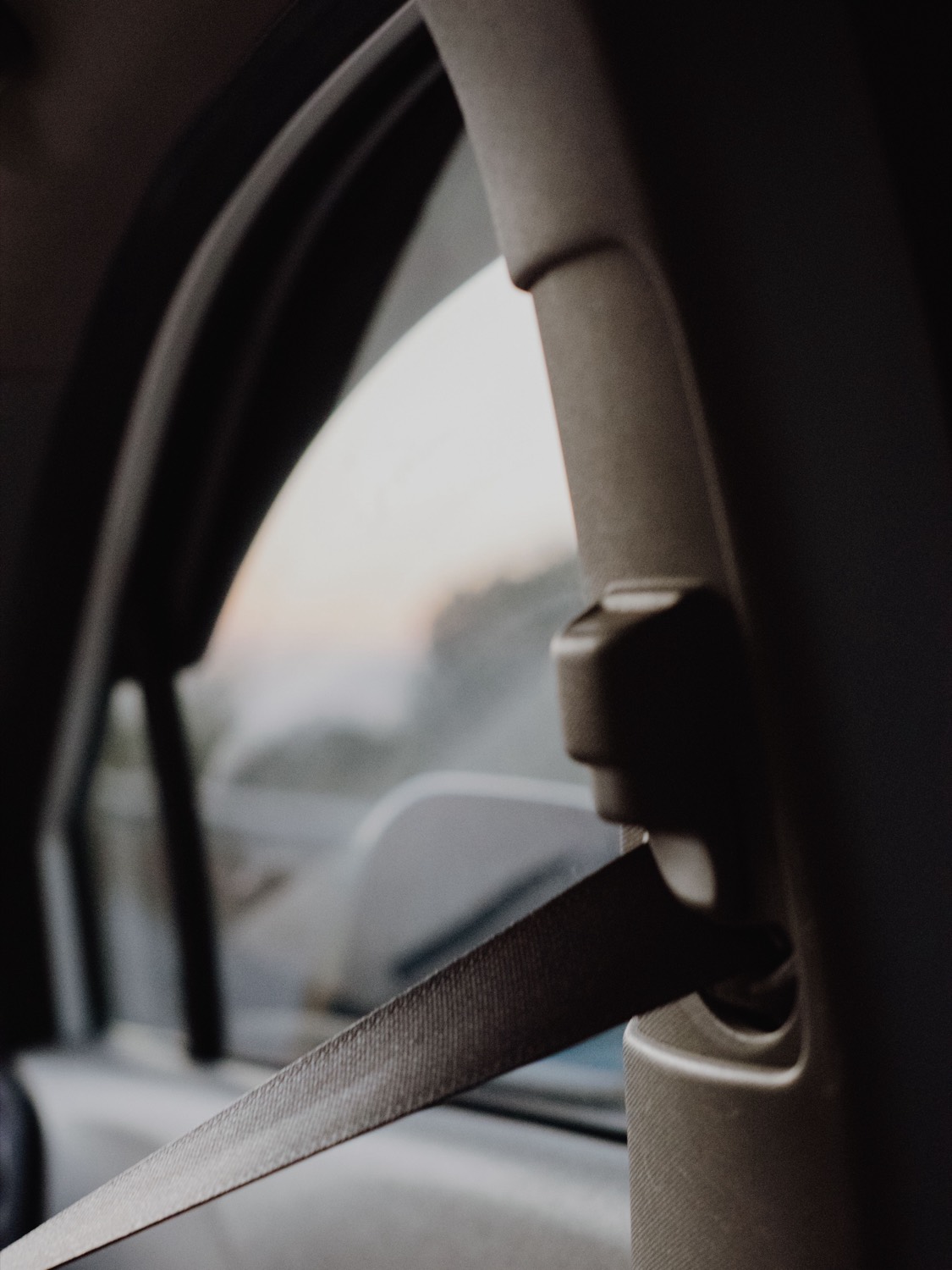 Upper seatbelt anchor and height adjuster on a vehicle B-pillar, photographed against evening light through the side window.