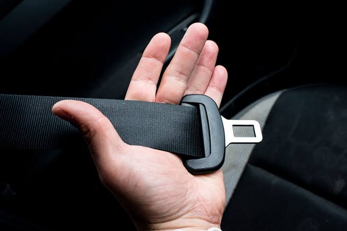 An open hand supporting a black seatbelt tongue plate and webbing inside a vehicle, showing the metal latch plate in detail.
