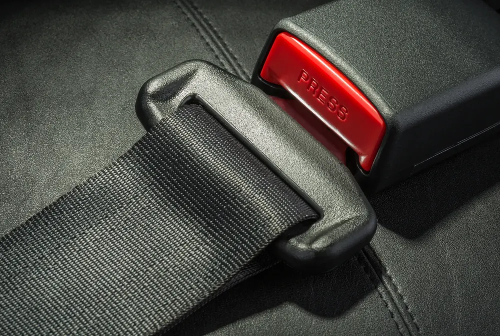 Close detail of seatbelt webbing, the moulded tongue plate and a buckle receiver marked PRESS on a black leather seat.