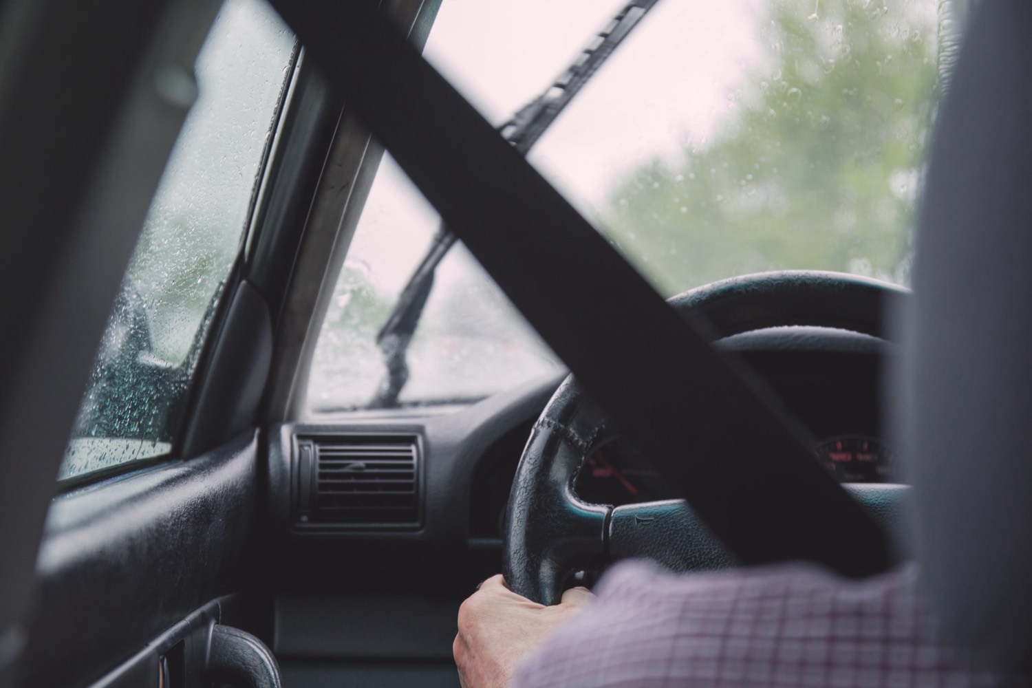 View from inside a vehicle cabin on a wet day, with the shoulder belt running across the driver towards the steering wheel.