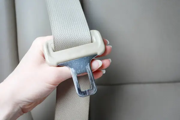 A hand holding a light-coloured seatbelt tongue plate, with the latch slot and webbing clearly visible for inspection.